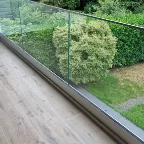 Balkon mit Glasgeländer und Holzoptik-Fliesen, Blick auf gepflegten Garten mit Hecke.
