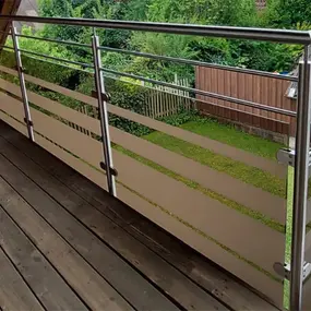 Balkongeländer aus Edelstahl mit Milchglas und Holzterrasse, moderner Gartenblick.
