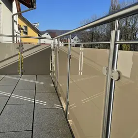 Moderner Balkon mit Edelstahlgeländer, Sichtschutz und grauen Bodenfliesen bei Sonnenschein.
