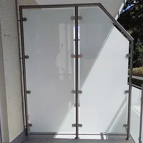 Balkon mit moderner Sichtschutzwand aus satiniertem Glas und Edelstahlrahmen bei Tageslicht.