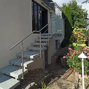 Außentreppe aus Granit mit Edelstahlgeländer vor einem Haus, umgeben von Blumen und Sträuchern.