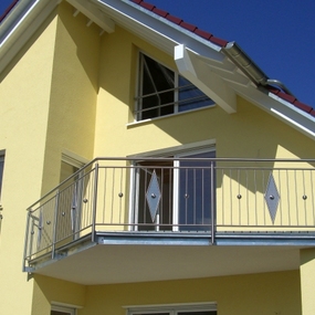 Modernes Einfamilienhaus mit gelber Fassade, Balkon und roten Dachziegeln bei Sonnenschein.